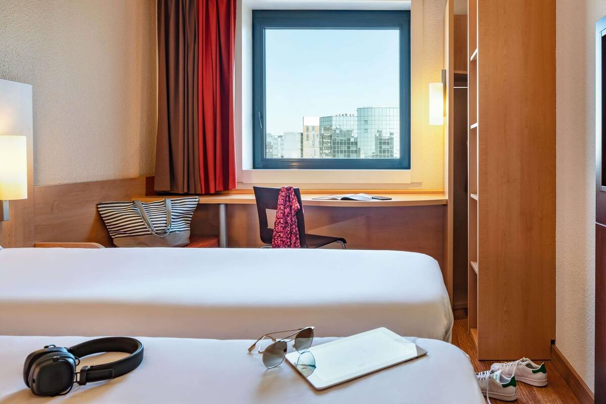 Hotelkamer met een bed, een bureau met stoel, een raam met uitzicht op gebouwen, en een paar koptelefoons en zonnebrillen op het bed.