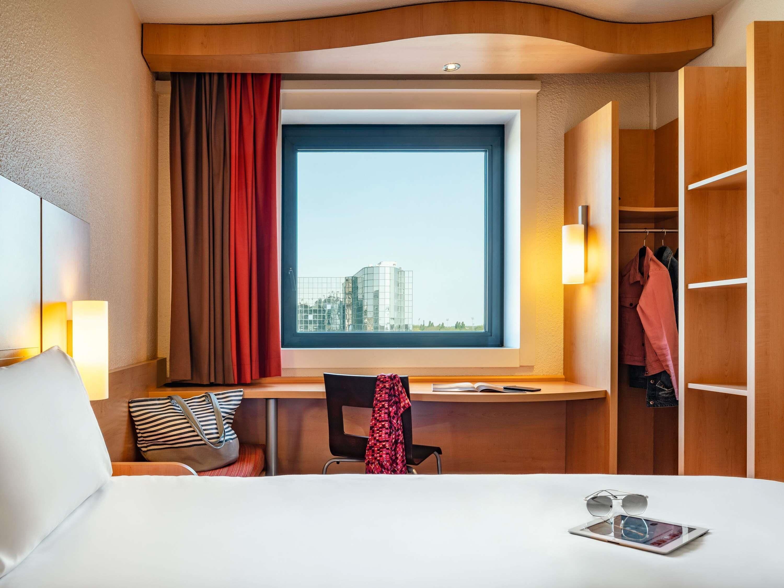 Ein Hotelzimmer mit Bett, Schreibtisch, Stuhl, Fenster mit Blick auf Gebäude und einem Kleiderschrank.