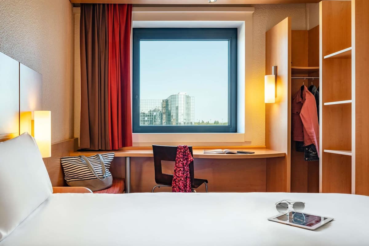 Ein Hotelzimmer mit Bett, Schreibtisch, Stuhl, Fenster mit Blick auf Gebäude und einem Kleiderschrank.