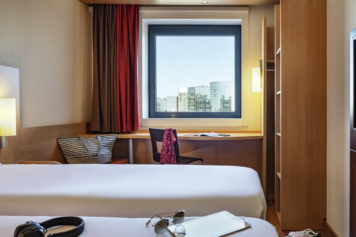 Une chambre d’hôtel avec deux lits, un bureau, une fenêtre donnant sur la ville, une lampe et une paire d’écouteurs posés sur le lit.