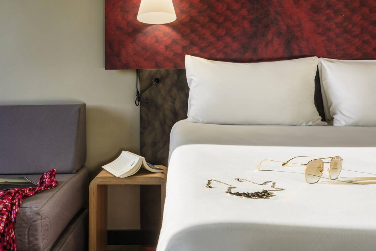 Une chambre d’hôtel avec un lit, une table de chevet, une lampe de lecture et une paire de lunettes.
