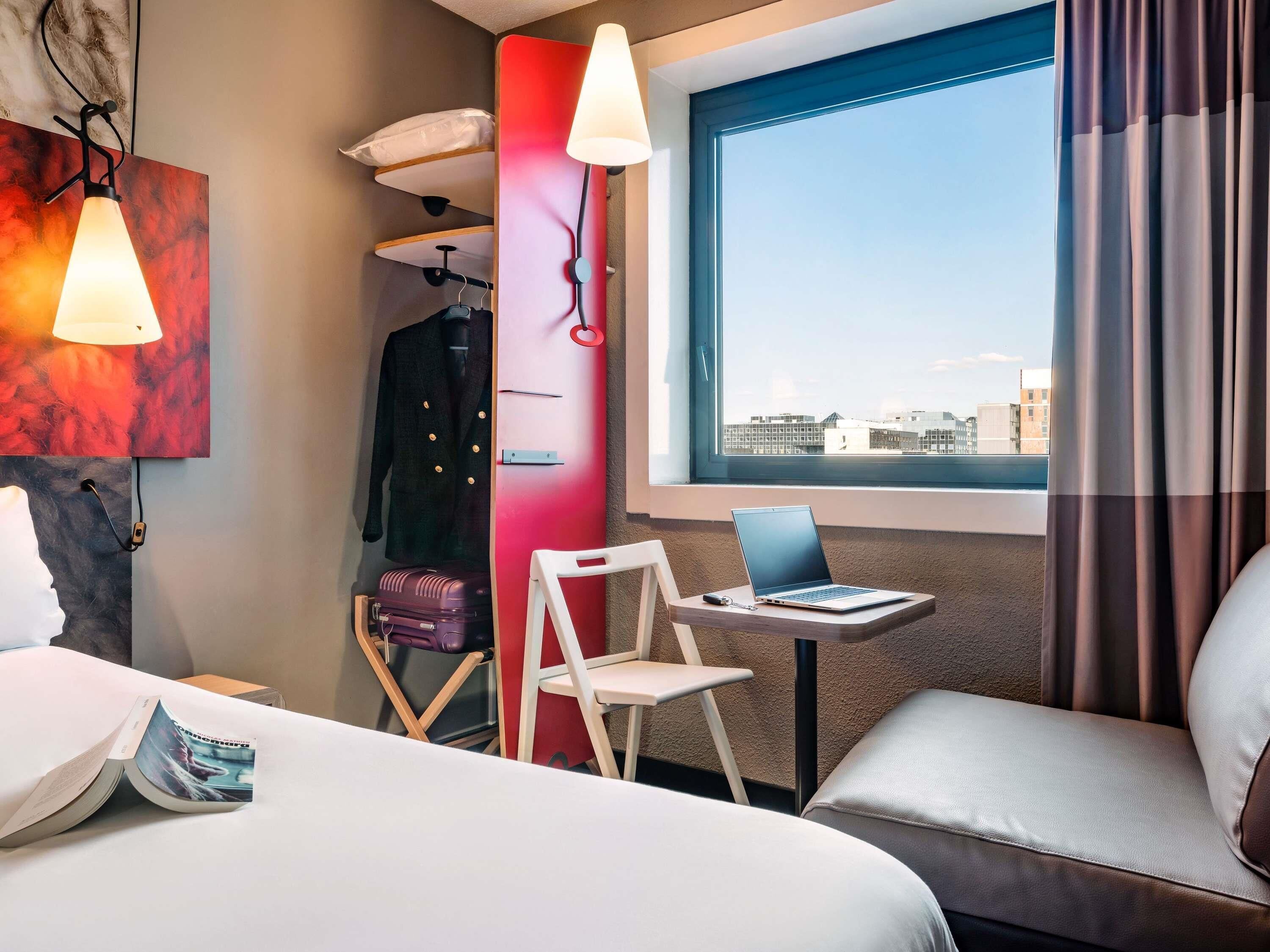 Habitación de hotel con cama, escritorio con una computadora portátil, silla, maleta y vista a la ciudad a través de una ventana.