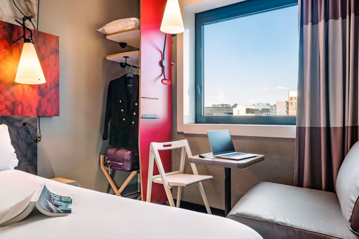 Une chambre d’hôtel avec un lit, un bureau avec un ordinateur portable, une chaise, une valise et une vue sur la ville par la fenêtre.