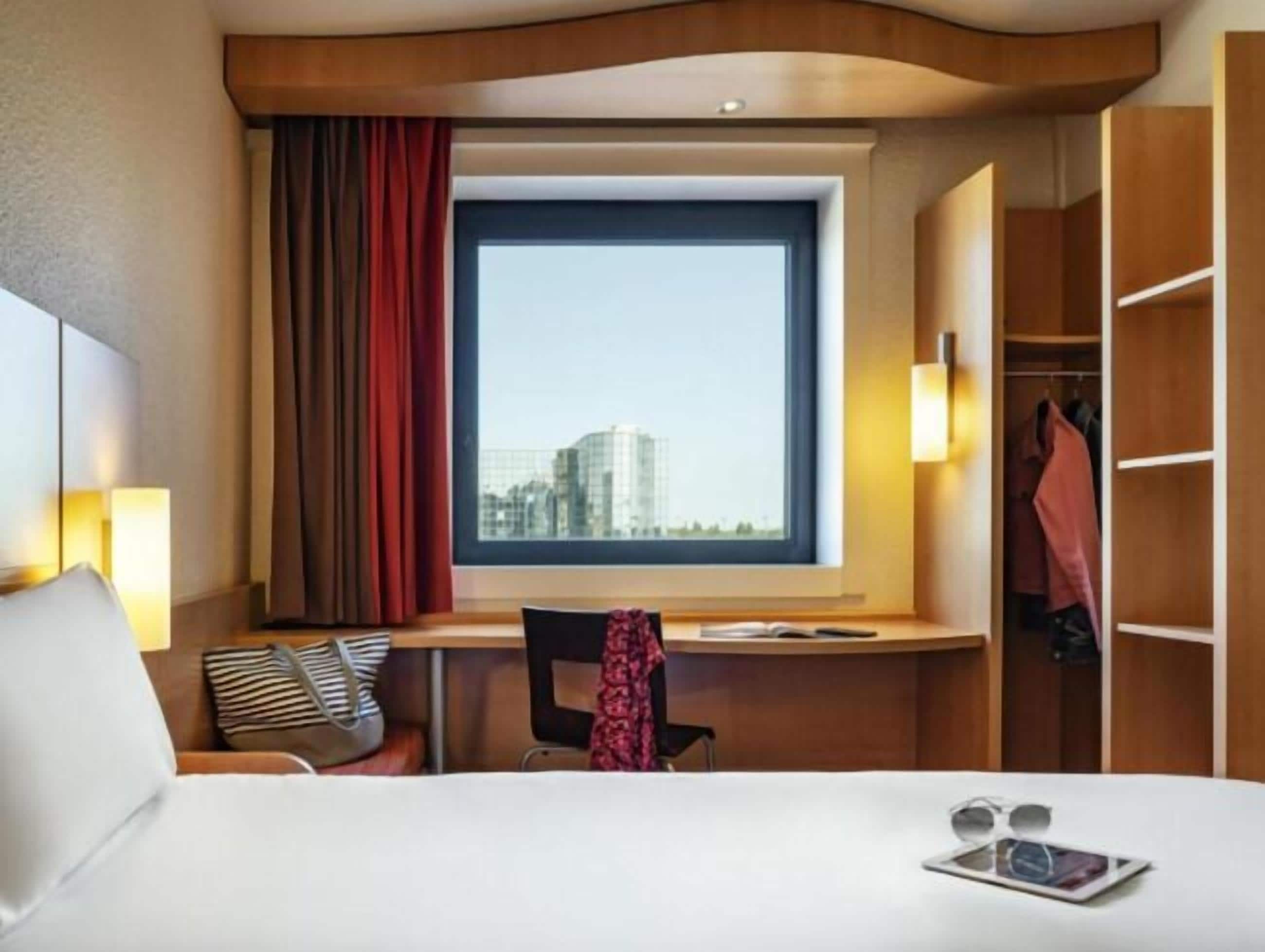 A hotel room with a bed, a desk with a chair, a window with a view of buildings, and a wardrobe.