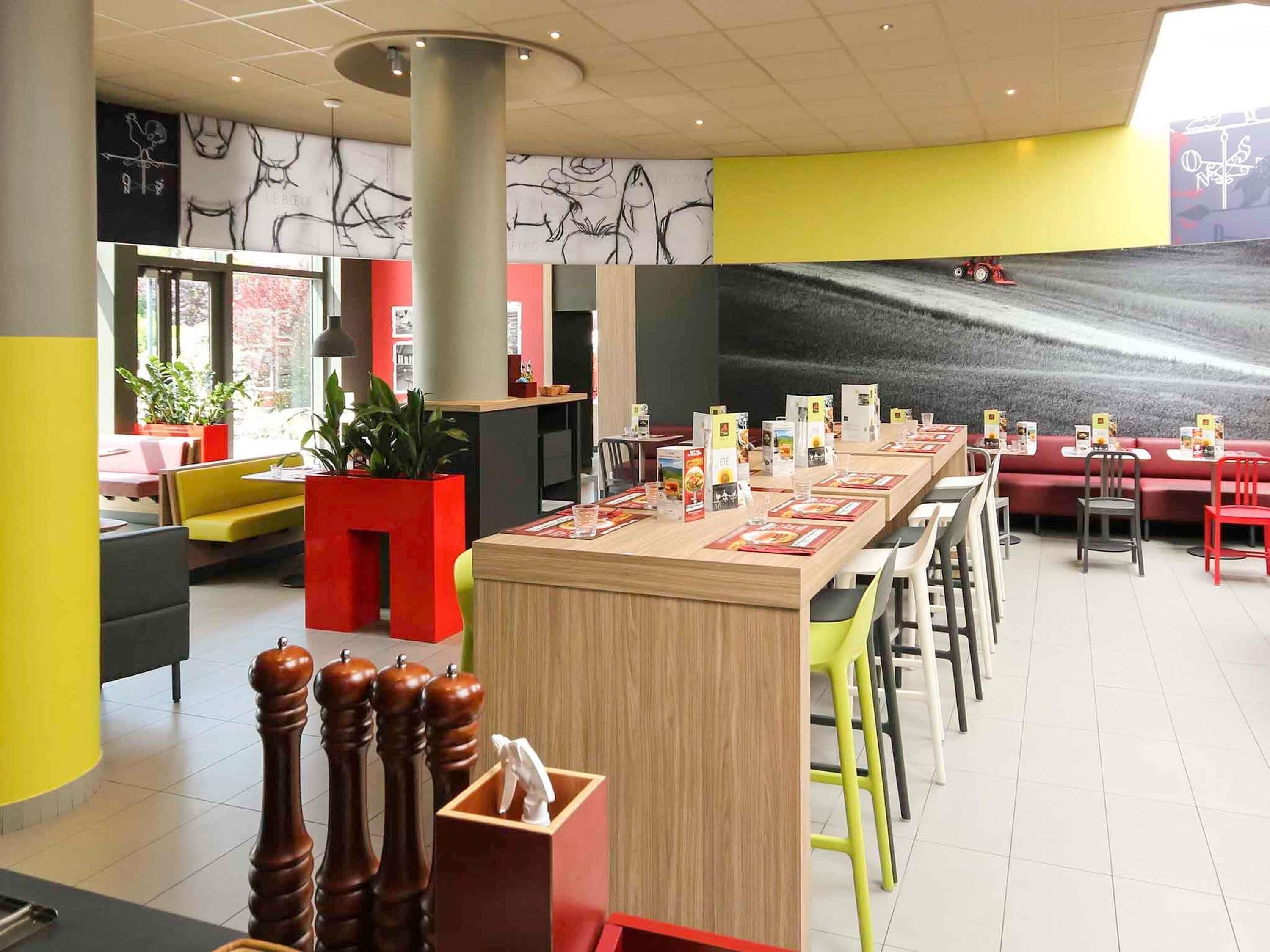 A modern restaurant with a long wooden table, red and yellow seating, and abstract wall art.