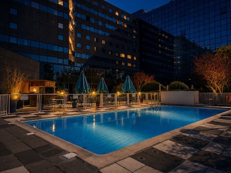 A swimming pool with lounge chairs and umbrellas, surrounded by a building with multiple windows and a checkered floor pattern.. ibis Marne La Vallée Noisy