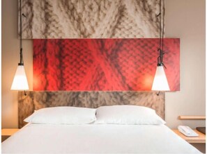 Une chambre d’hôtel avec un lit, deux lampes suspendues et un grand tableau abstrait au-dessus du lit.