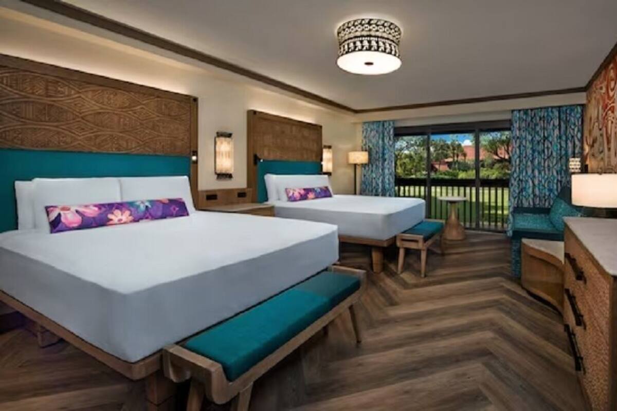A hotel room with two beds, a desk, a bench, and a view of the outdoors.