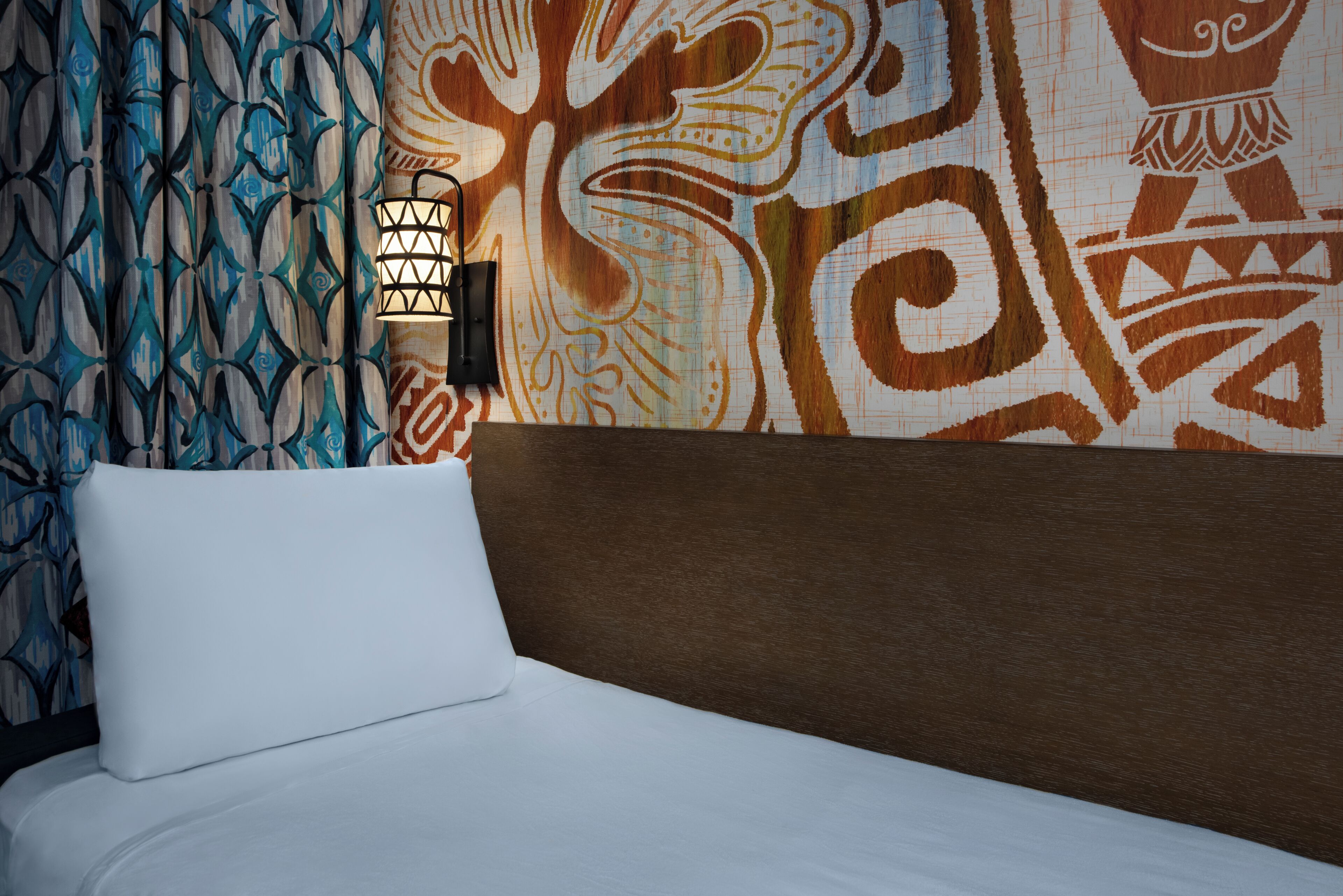 A bed with a white pillow and a brown headboard in a room with patterned wallpaper and a wall-mounted light fixture.