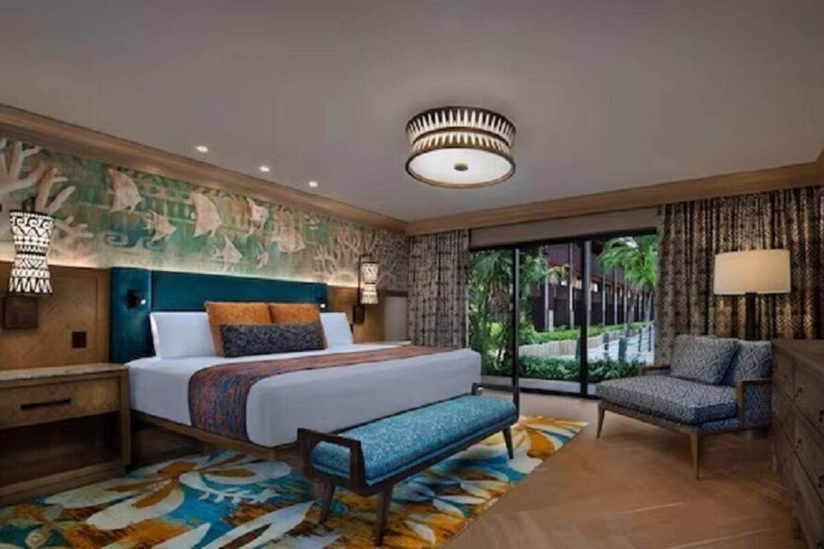 A hotel room with a bed, a bench, a chair, a dresser, a lamp, a ceiling light, and a view of greenery outside.