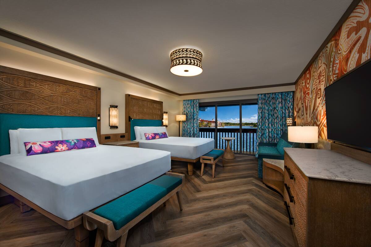Habitación de hotel con dos camas, un escritorio, una silla, un balcón con vistas y un televisor grande.