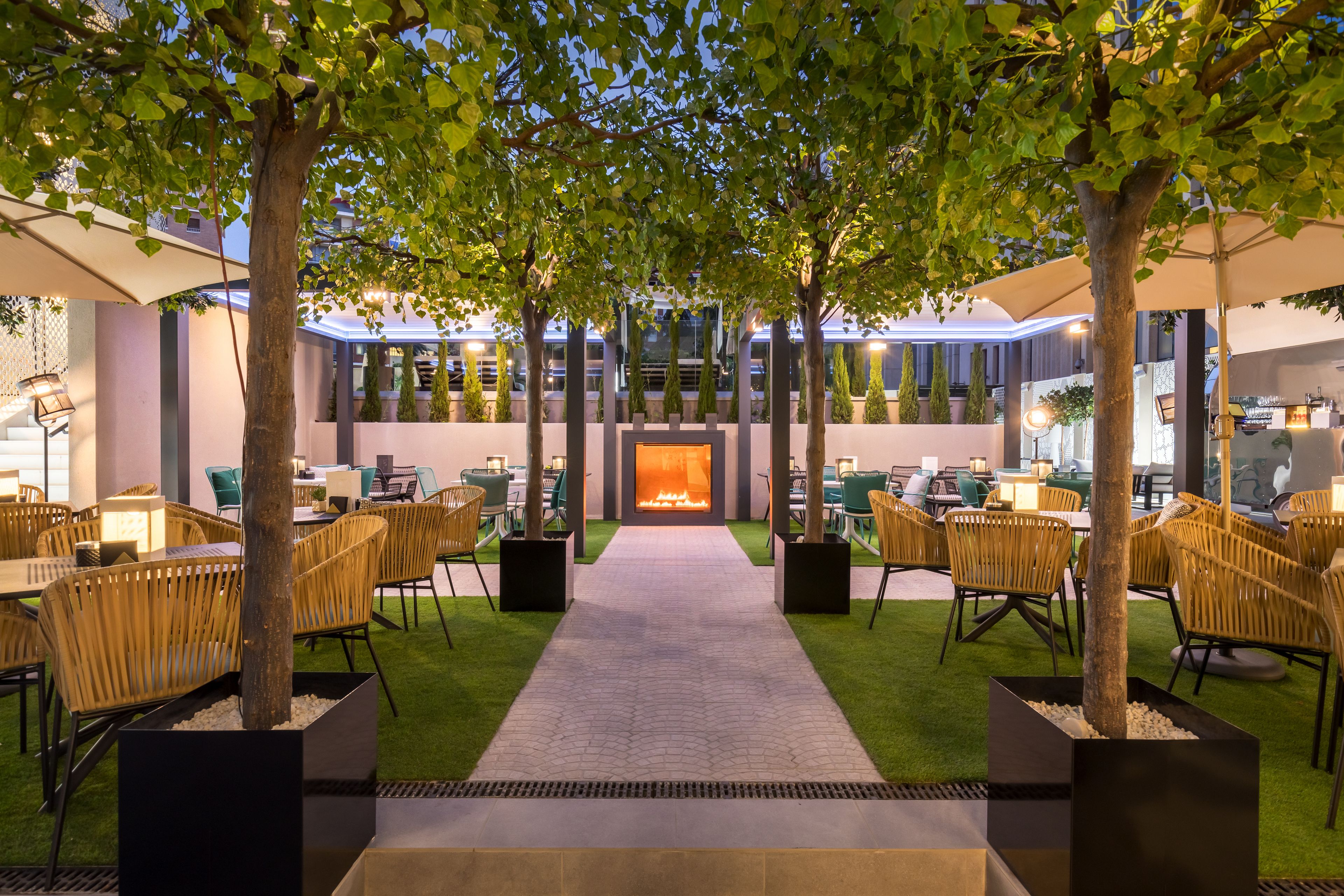 An indoor courtyard with a central fire feature, surrounded by tables and chairs, and trees providing shade.
