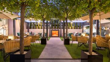 An indoor courtyard with a central fire feature, surrounded by tables and chairs, and trees providing shade.