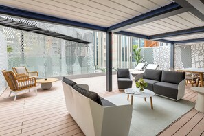 A modern outdoor living area with a wooden floor, a white sofa, a grey sofa, a wicker chair, and a round table.