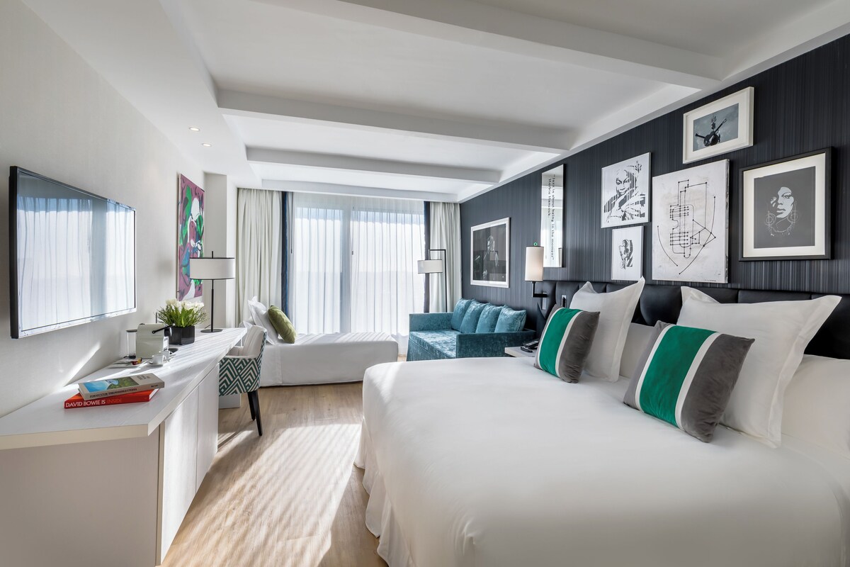 Una camera d'albergo moderna con un letto grande, una scrivania con un computer, una sedia e un divano. Alle pareti ci sono quadri incorniciati e una finestra con tende.