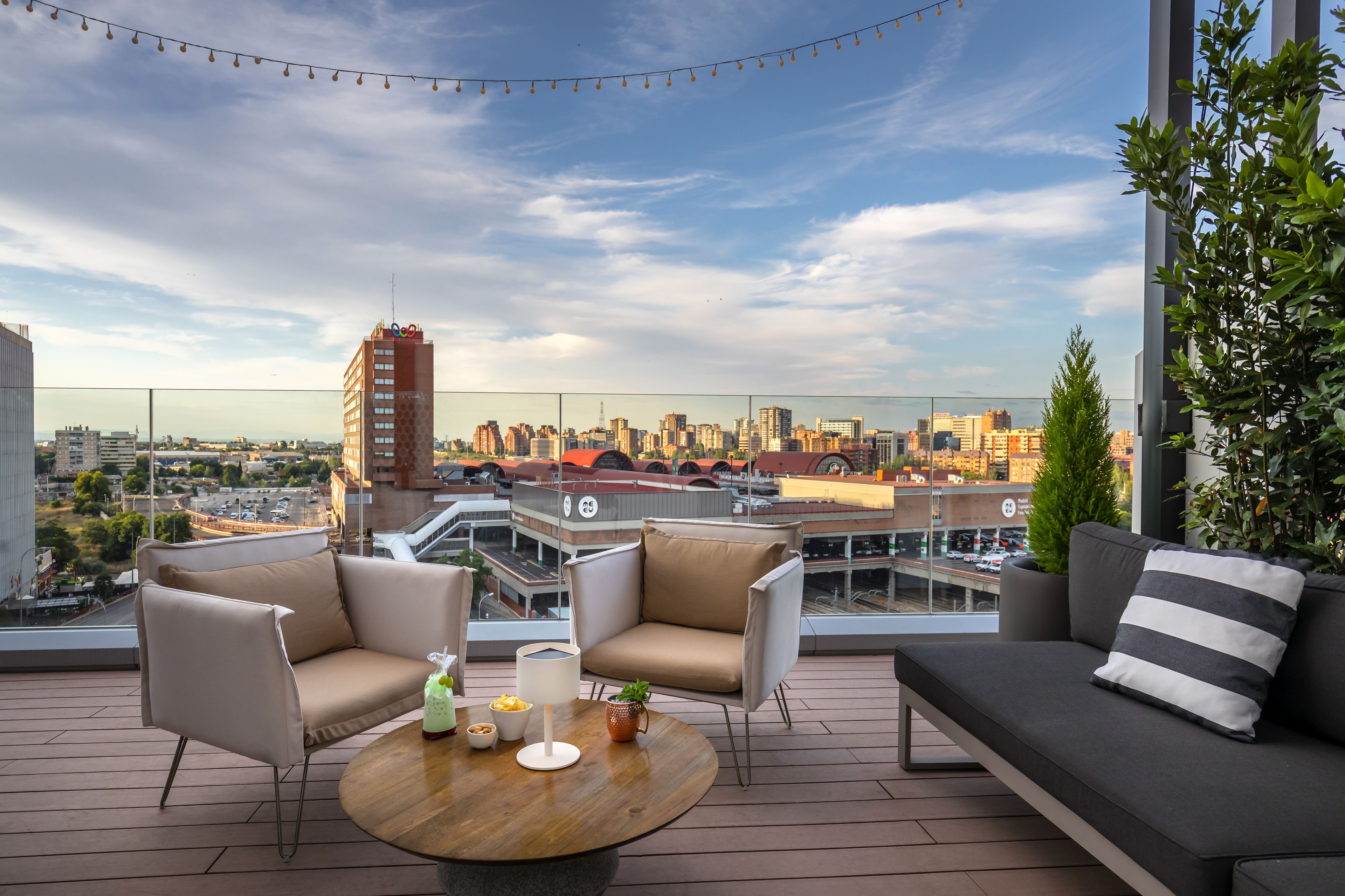A rooftop terrace with a city view, furnished with a sofa, chairs, and a round table with drinks and snacks.