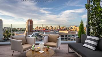 A rooftop terrace with a city view, furnished with a sofa, chairs, and a round table with drinks and snacks.