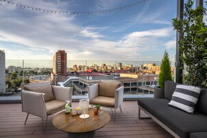 A rooftop terrace with a city view, furnished with a sofa, chairs, and a round table with drinks and snacks.