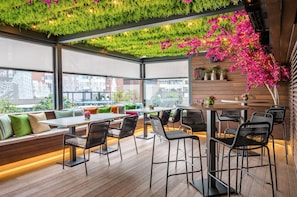 Un bar en la azotea con piso de madera, sillas de metal negro y una zona de estar con cojines.