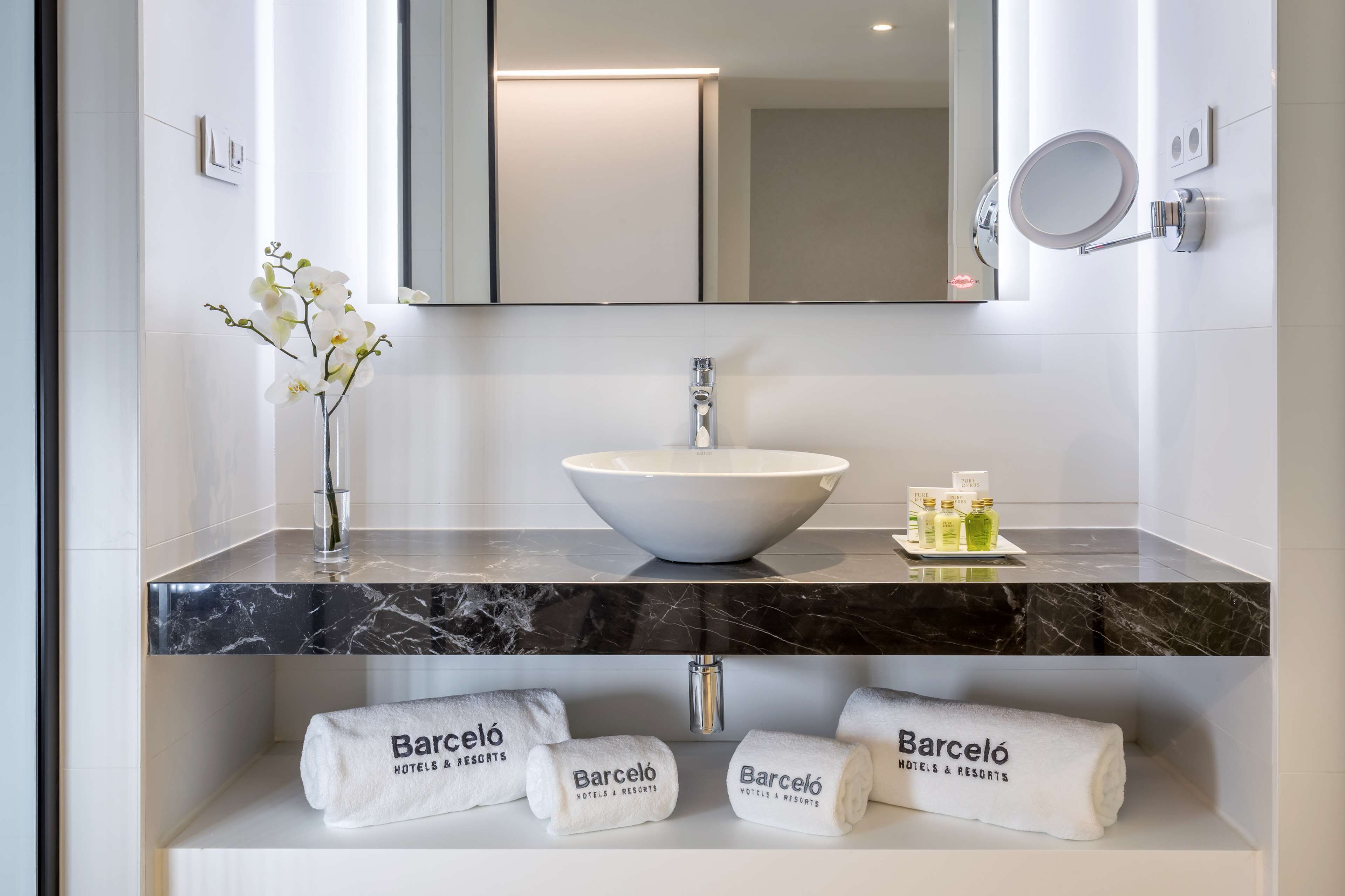 A modern bathroom with a white sink, a large mirror, and a vase with white flowers. There are towels and toiletries on the counter.