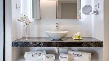 Un baño moderno con un lavabo blanco, un espejo grande y un florero con flores blancas. Hay toallas y artículos de aseo en la encimera.