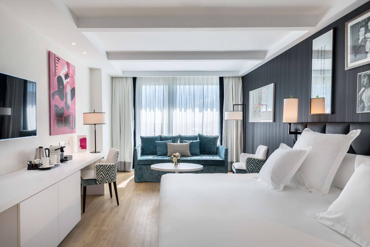A modern hotel room with a large bed, a sofa, a desk with a chair, a round coffee table, and a TV.