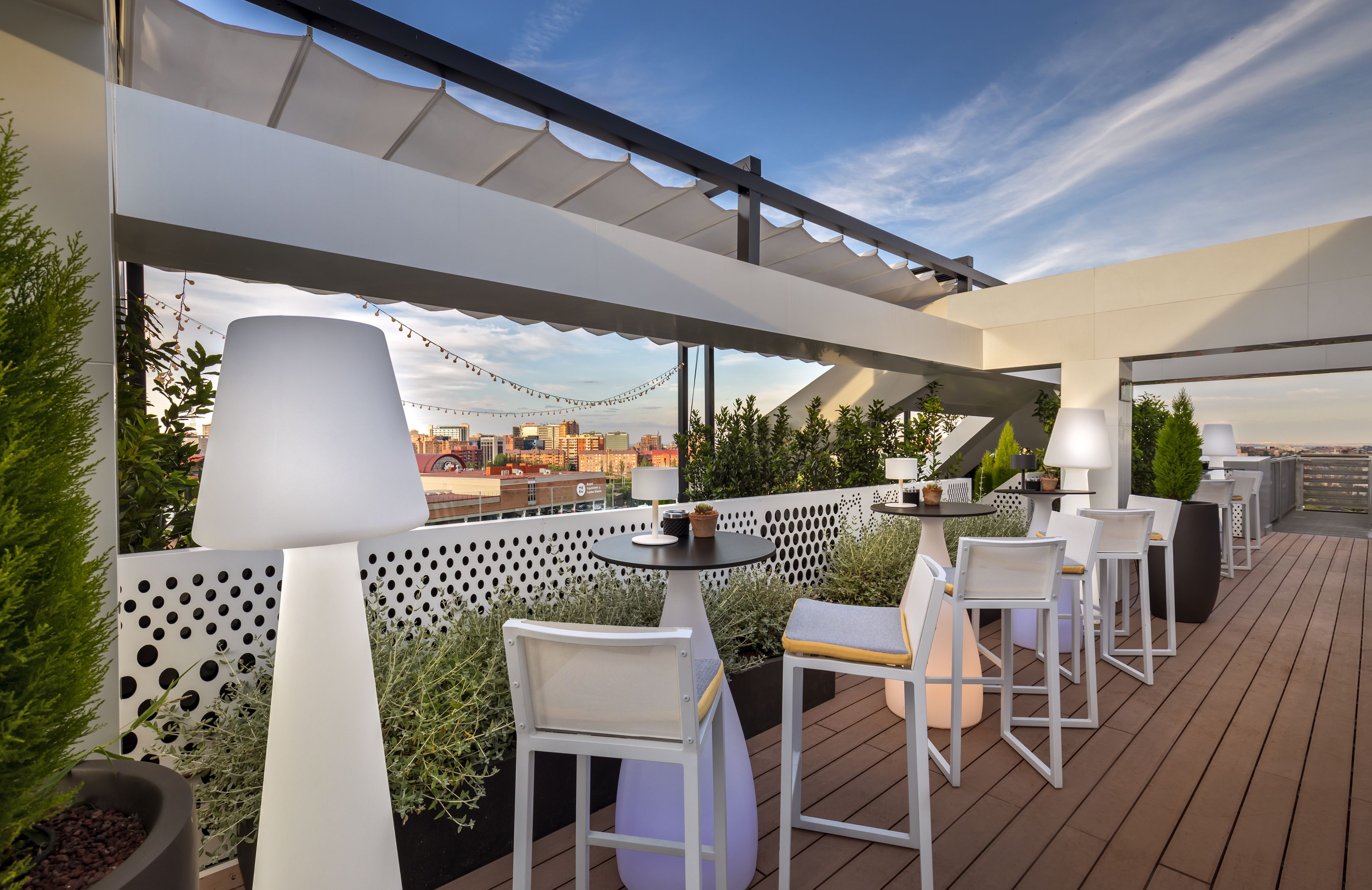 An outdoor patio with white chairs and tables, string lights, and a city view.