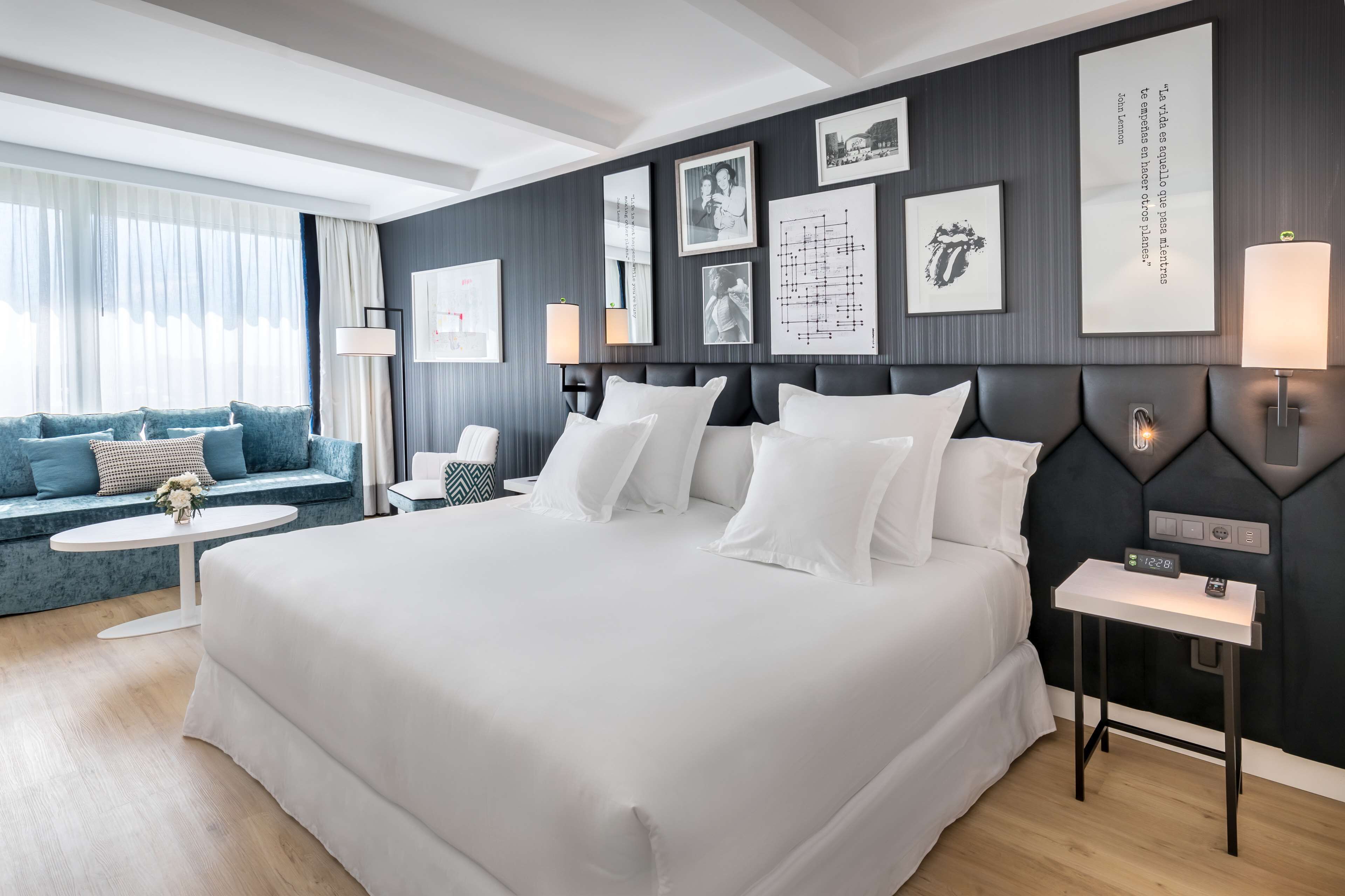 Una habitación de hotel moderna con una cama grande, un sofá, una mesita pequeña y una mesita de noche con una lámpara.