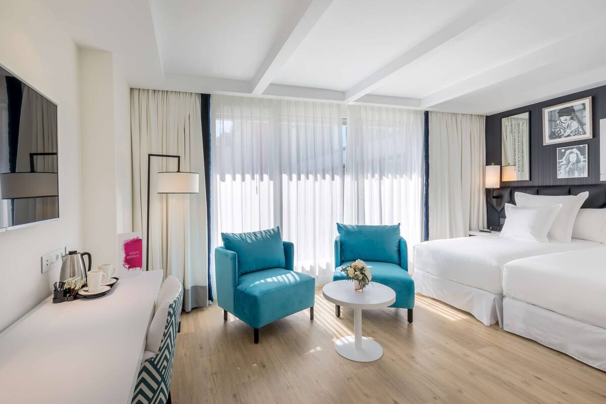A modern hotel room with a bed, two blue armchairs, a white sofa, a coffee table, and a TV on the wall.