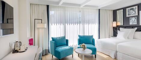 Una habitación de hotel moderna con una cama, dos sillones azules, un sofá blanco, una mesa de centro y un televisor en la pared.