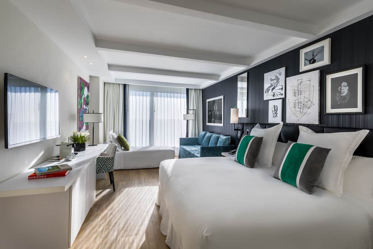 A modern hotel room with a large bed, a desk with a computer, a chair, and a sofa. There are framed pictures on the wall and a window with curtains.