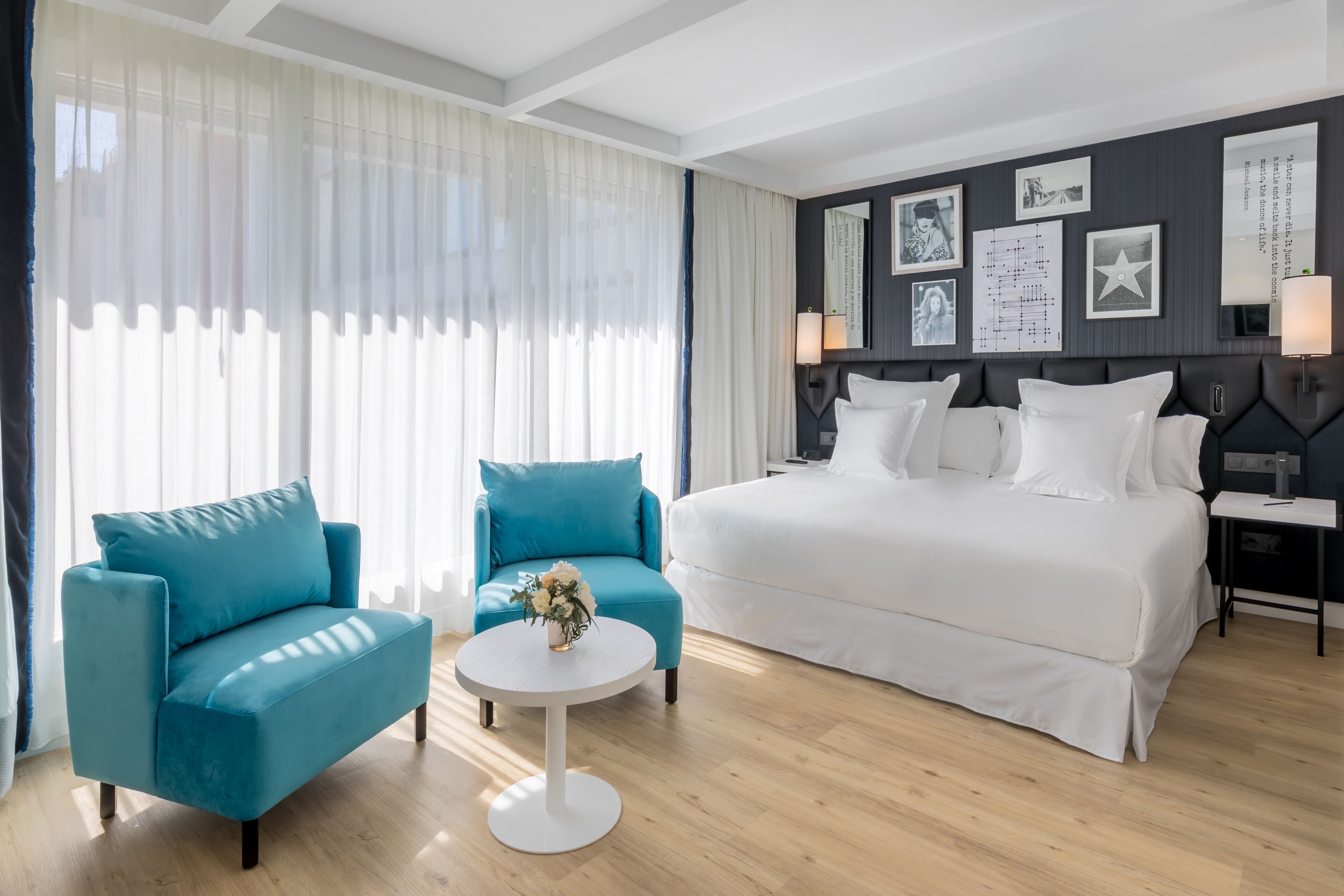 Ein Hotelzimmer mit einem großen Bett, zwei Sesseln, einem kleinen runden Tisch mit einer Vase und Blick auf die Stadt durch durchsichtige Vorhänge.