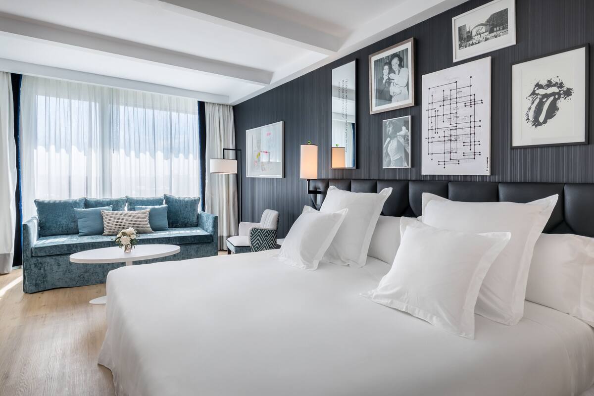 A modern hotel room with a large bed, a sofa, a round table, and a view of the outdoors.
