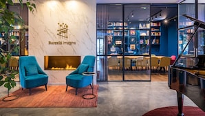 A modern lounge area with a fireplace, two teal armchairs, a grand piano, and a bookshelf filled with books. - Barceló Imagine (Madrid)