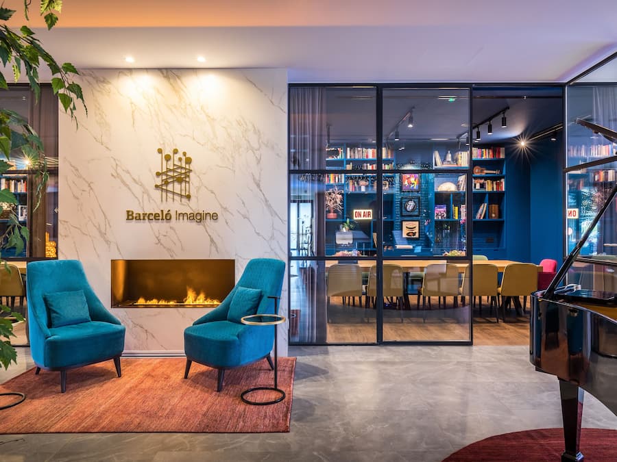 A modern lounge area with a fireplace, two teal armchairs, a grand piano, and a bookshelf filled with books.