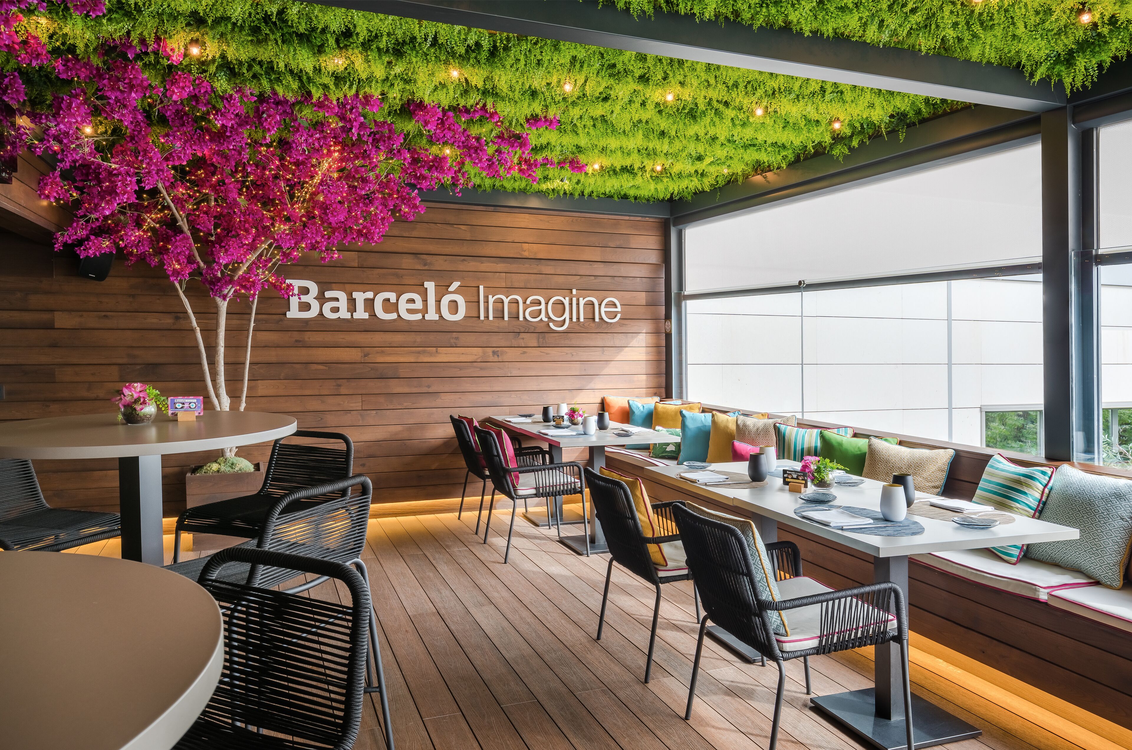 Una zona de estar moderna al aire libre con piso de madera, sillas de metal negro y una pared de madera con el texto "Barceló Imagine".