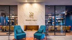 A modern lounge area with a fireplace, two blue armchairs, and a glass wall displaying a library. - Barceló Imagine (Madrid)