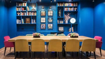 Una habitación con una larga mesa de madera rodeada de seis sillas, una estantería llena de libros y objetos decorativos, y una pared de ladrillos azules.