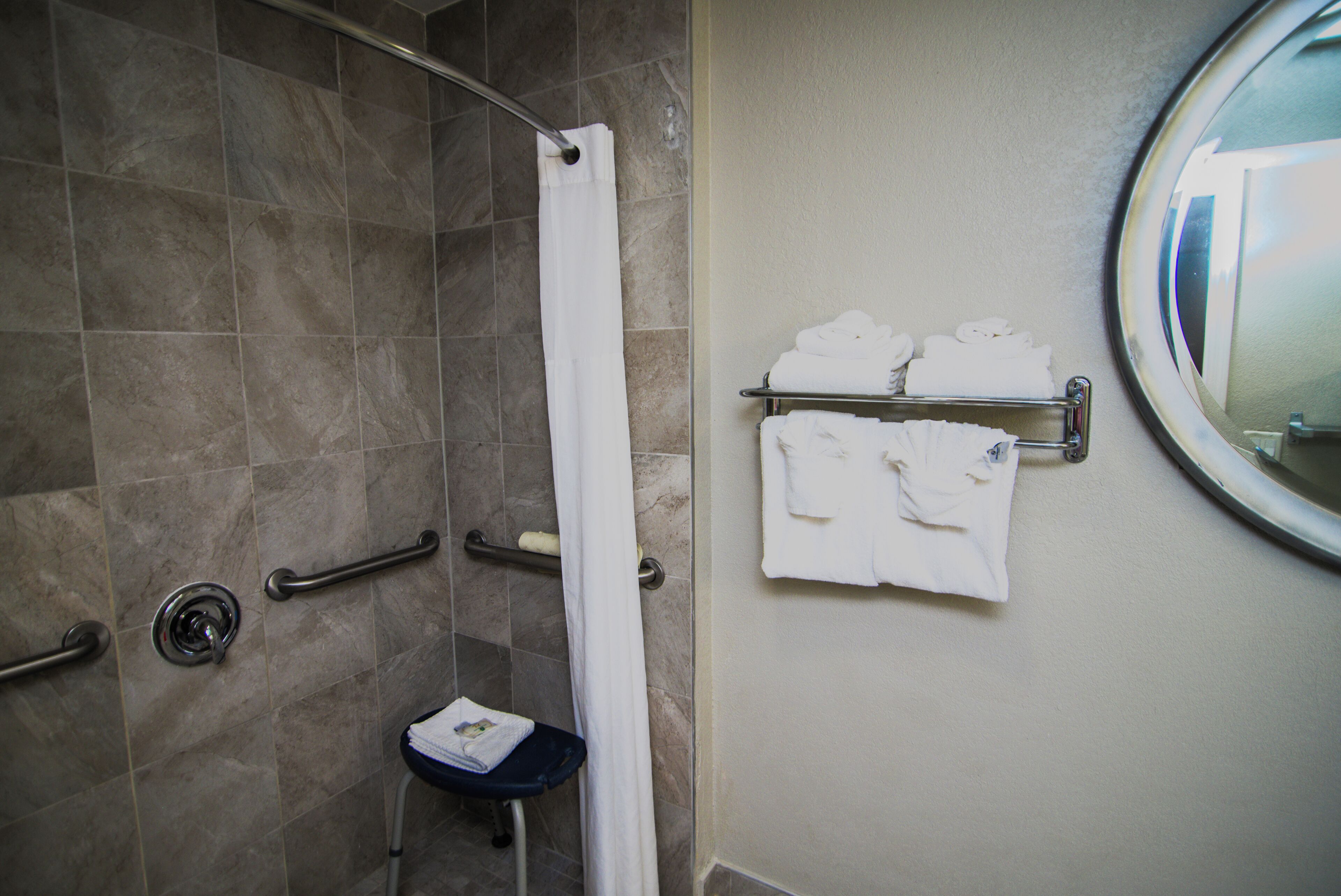 Un baño con ducha, barras de apoyo, un perchero con toallas dobladas y un espejo redondo.
