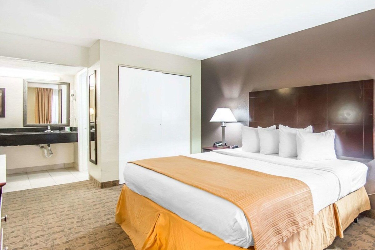 A hotel room with a bed, a nightstand, a lamp, a mirror, and a bathroom with a sink and toilet.