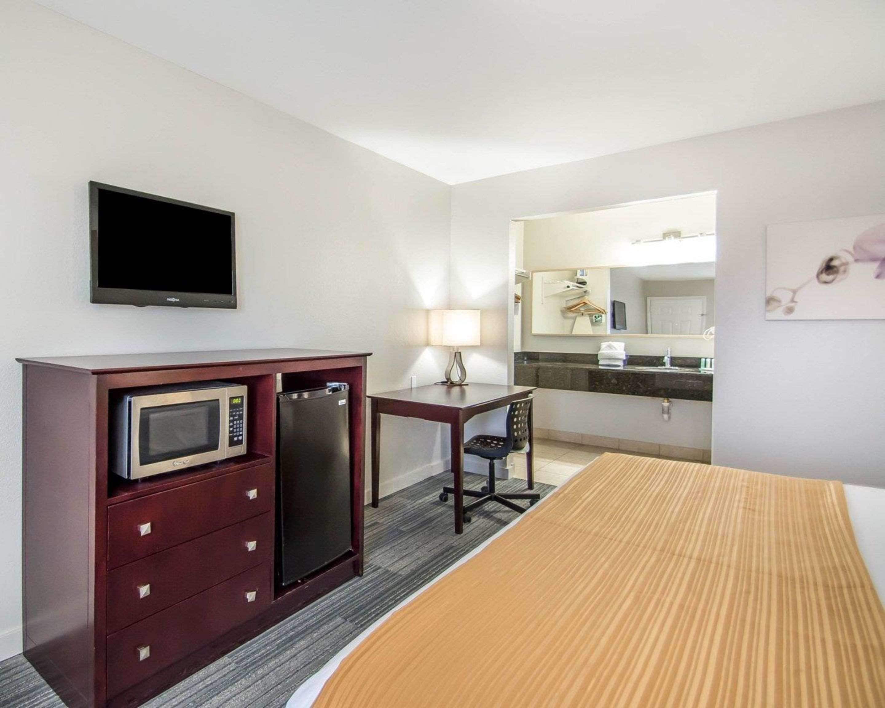 A hotel room with a bed, a desk with a microwave, a refrigerator, a chair, a TV, a bathroom with a mirror, and a towel rack.