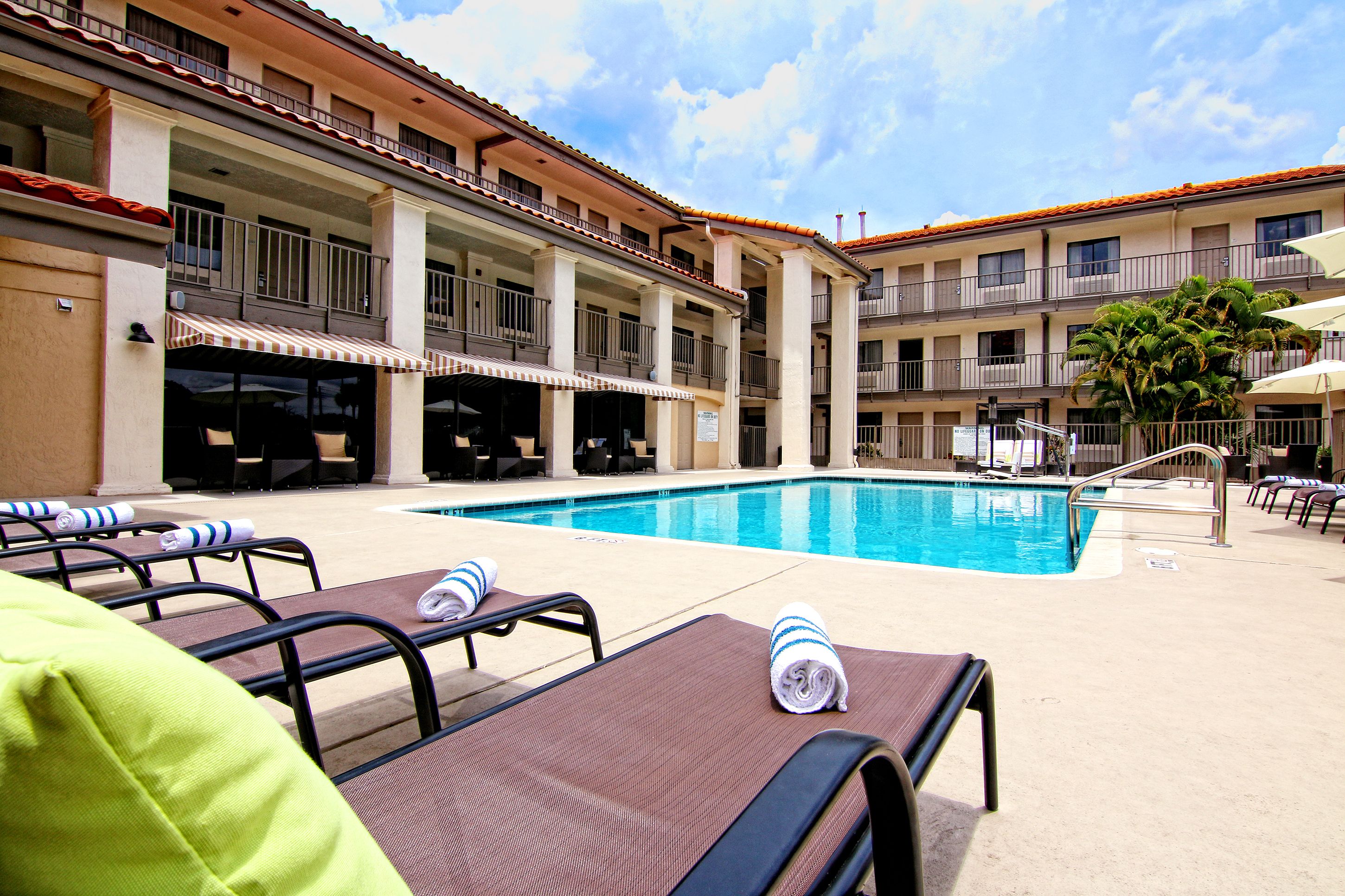 Un espace piscine doté de chaises longues et de parasols, une piscine à l’eau d’un bleu limpide et un immeuble à plusieurs étages avec des balcons.