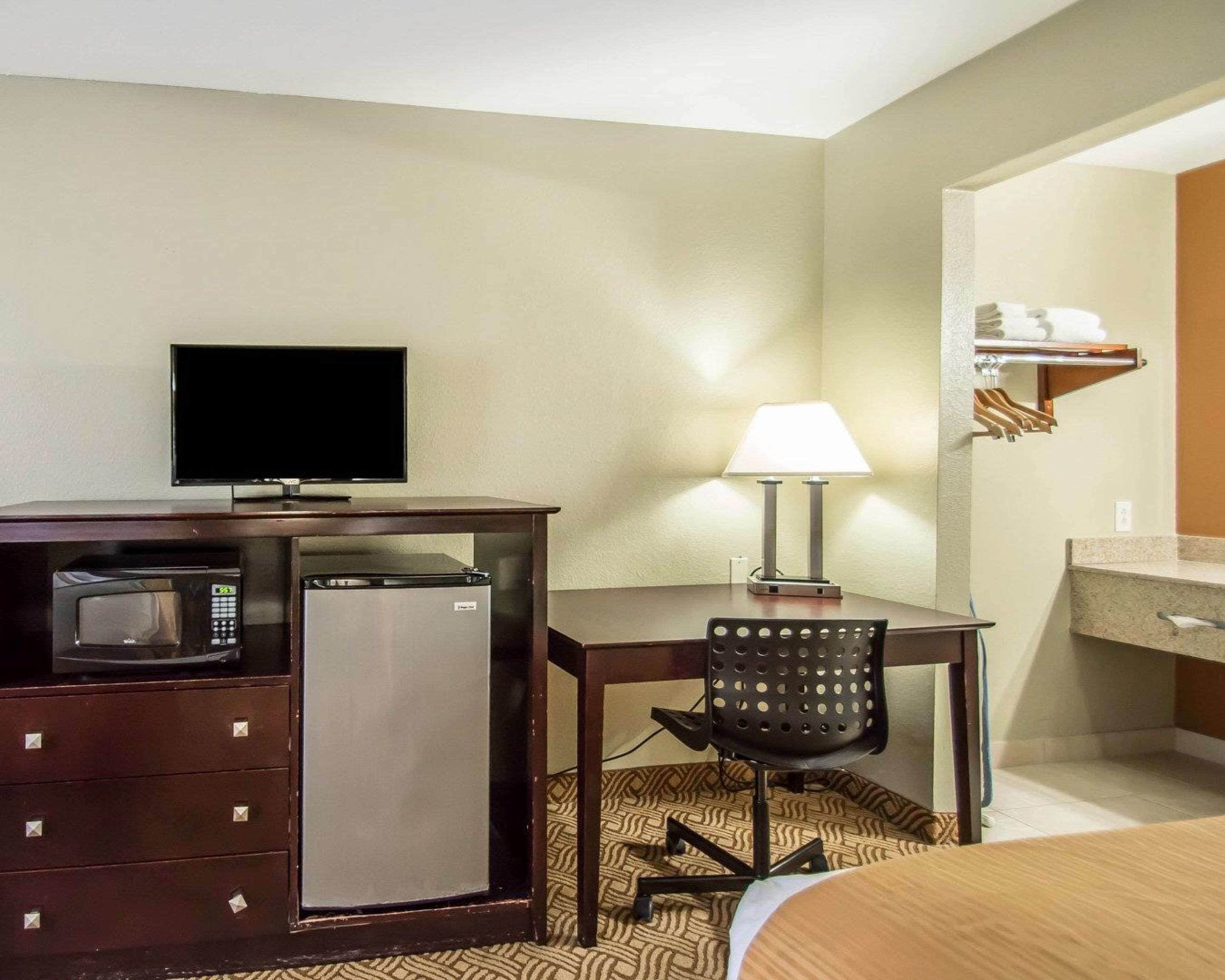 Habitación de hotel compacta con escritorio, televisión, microondas, refrigerador, silla, lámpara y cama.