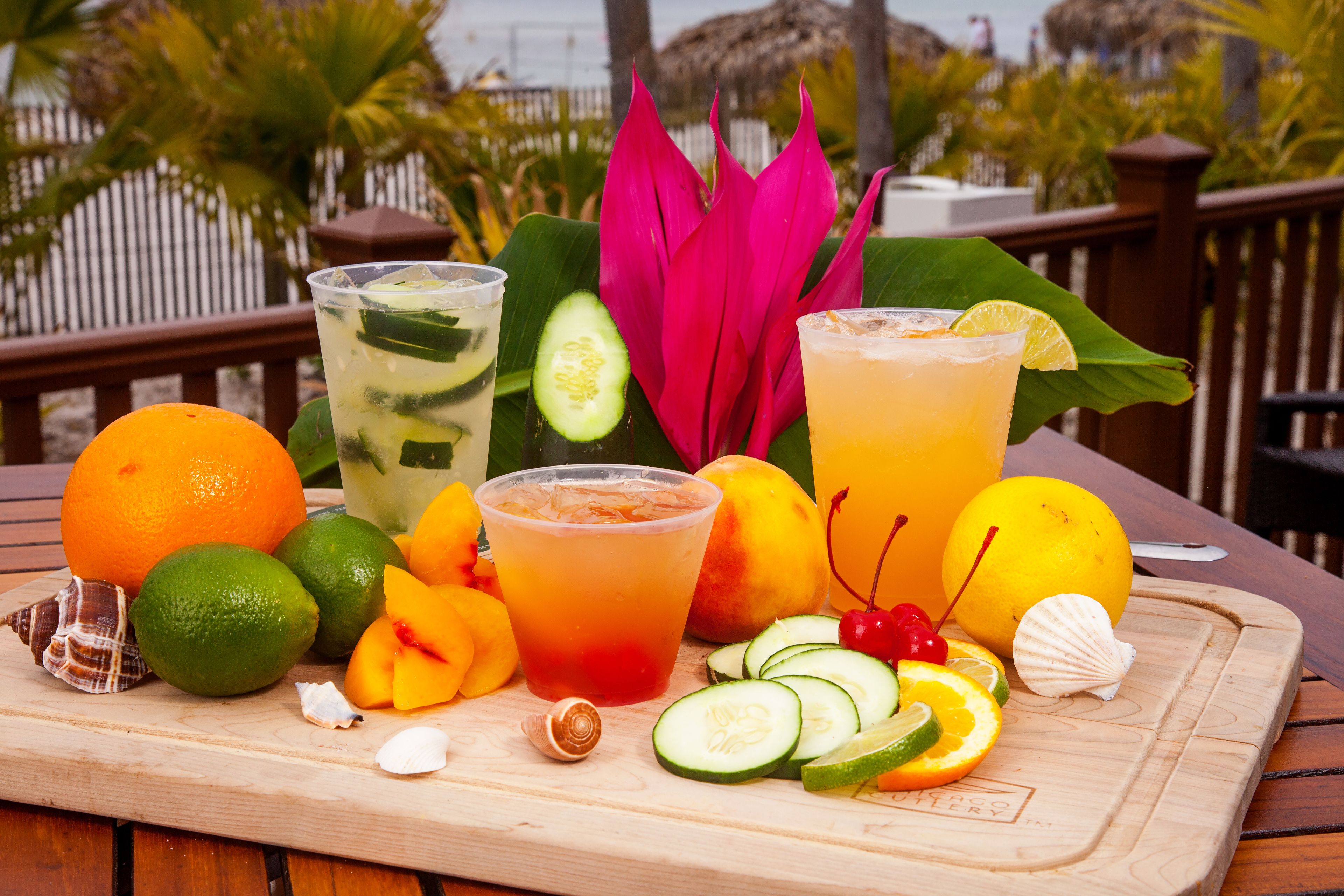 Uma mesa de madeira com diversas frutas e bebidas, incluindo laranjas, limões e uma abacaxi, com vista para um deck de madeira e palmeiras.