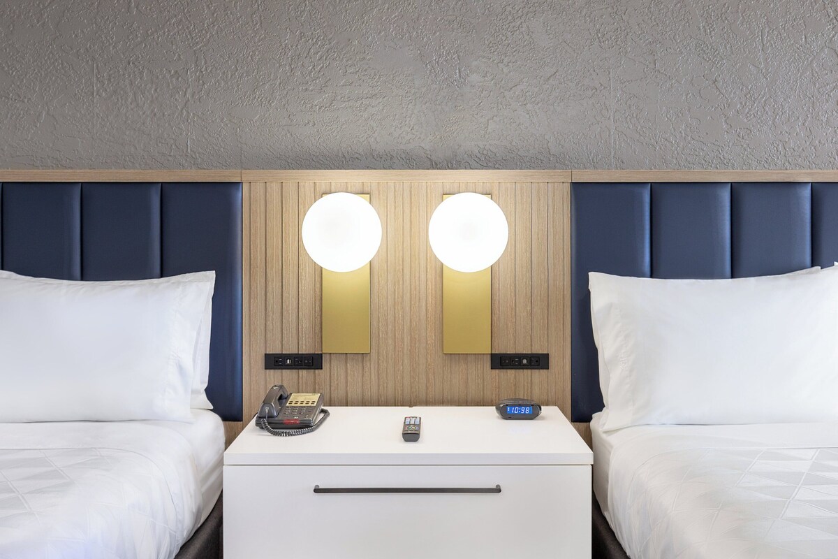 Habitación de hotel con dos camas, cabecero de madera, mesita de noche blanca con teléfono y reloj, y una lámpara de pared.
