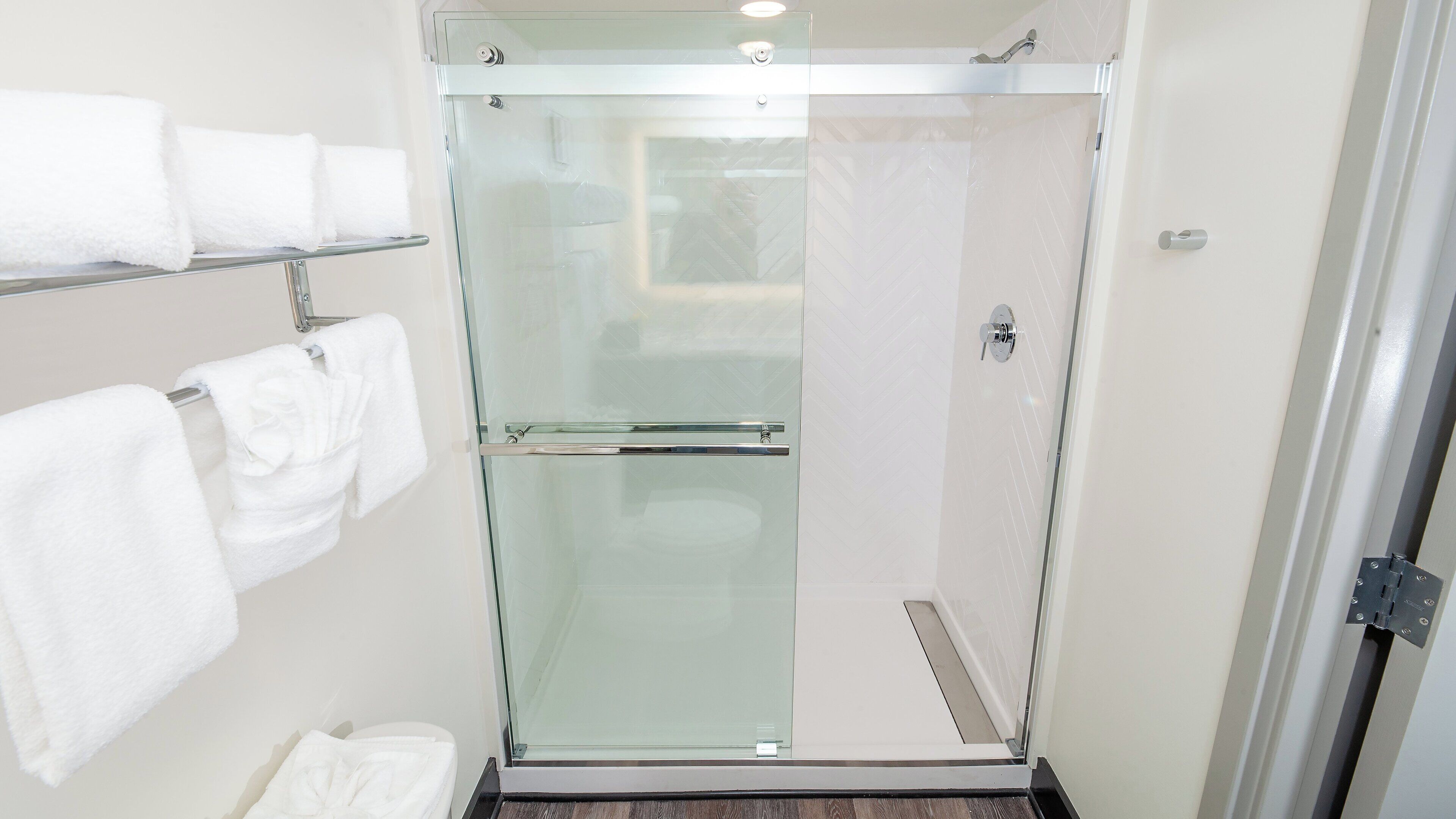 A glass-enclosed shower stall with a white tiled wall and a towel rack with folded towels.