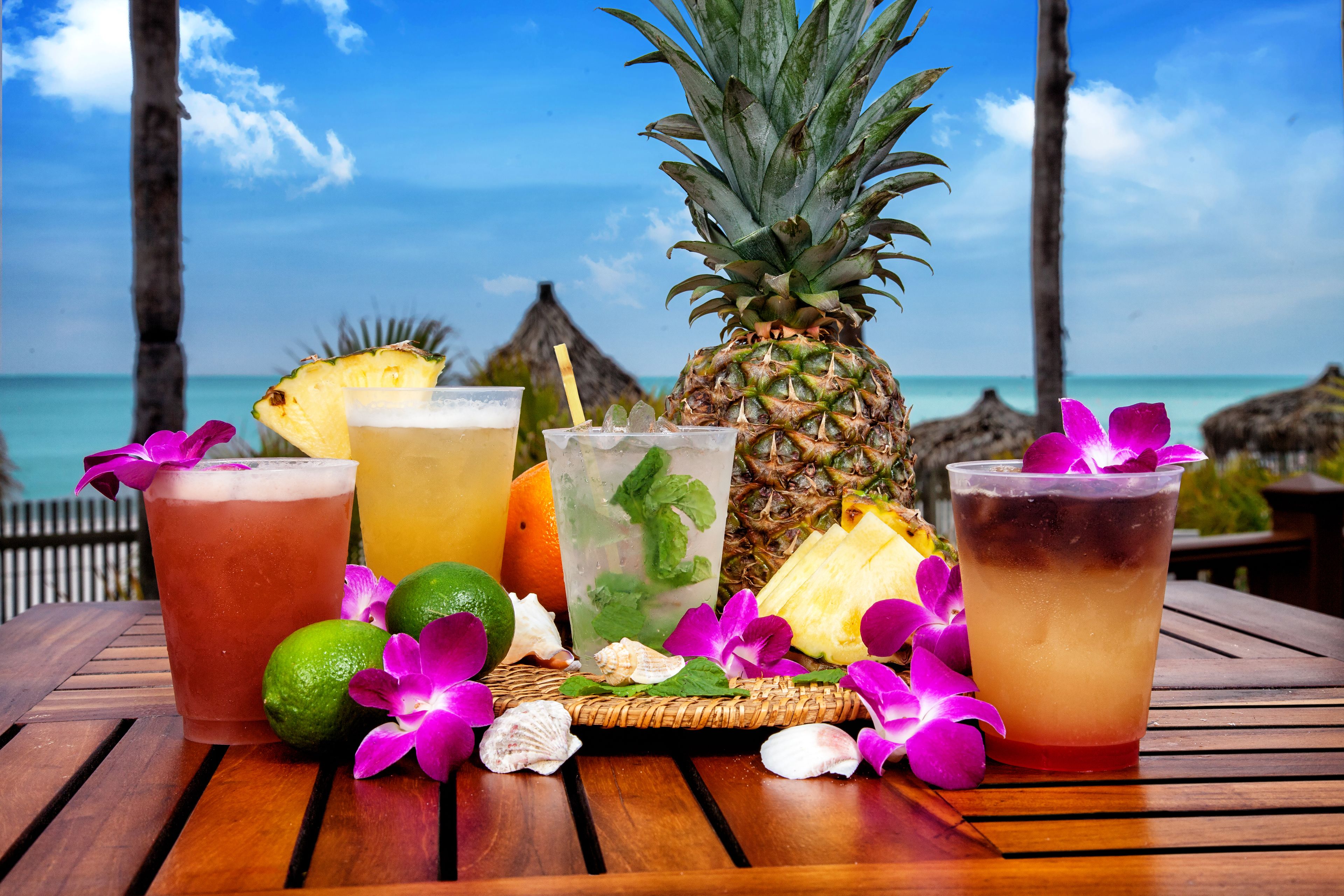 A wooden table with drinks and tropical fruits, set against a beach backdrop.