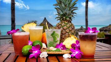 A wooden table with drinks and tropical fruits, set against a beach backdrop.