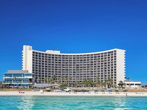 Een groot, meerhoog hotel met een gebogen gevel, direct naast een strand met helderblauw water.