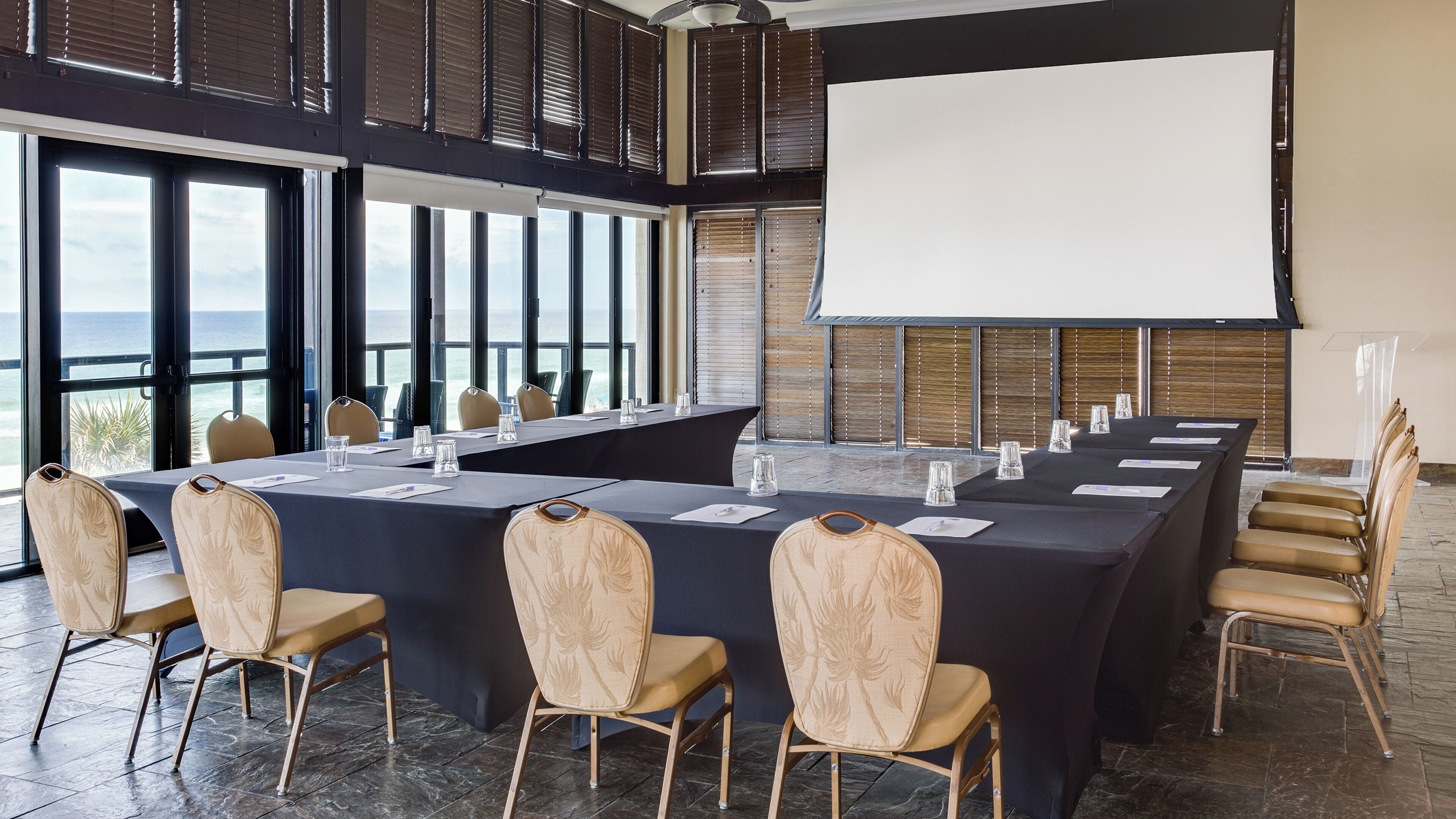 A conference room with a long table, chairs, a projector screen, and large windows overlooking a body of water.